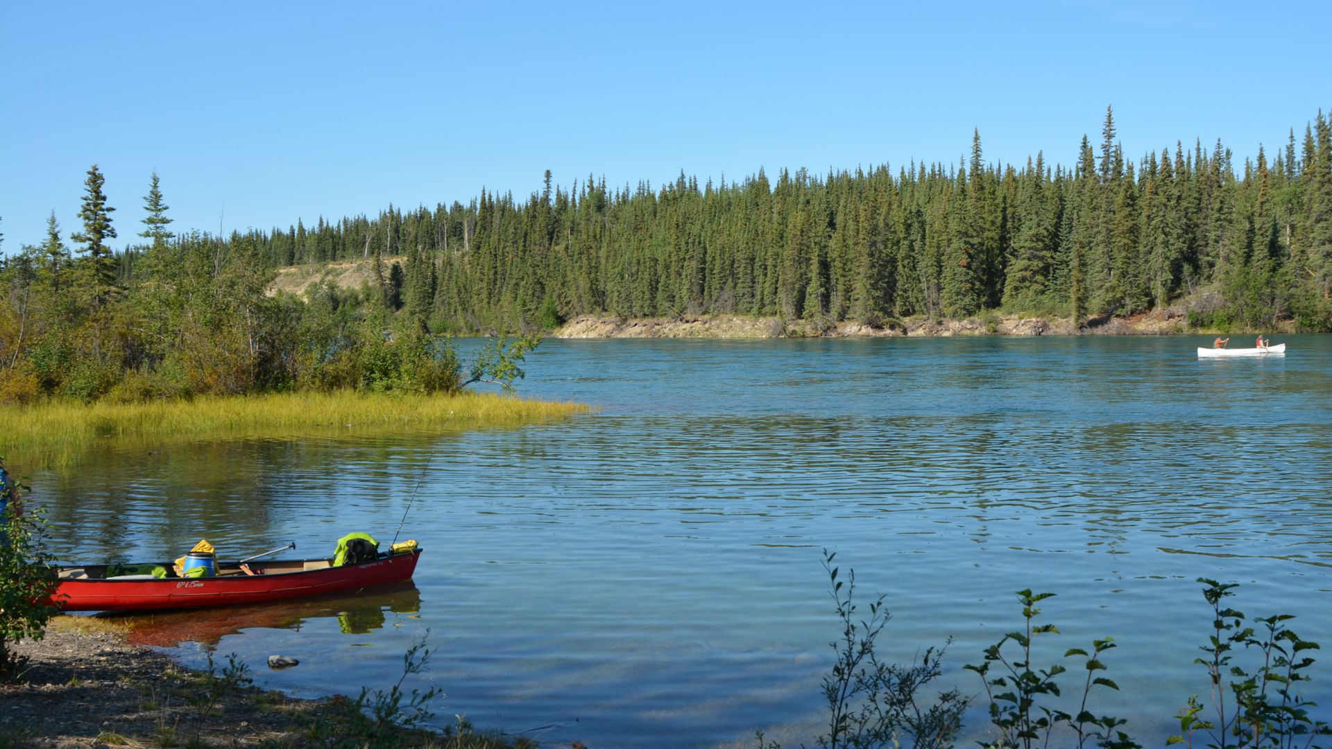 A canoe rests on the shore of the Thirty Mile River on a sunny day, while a canoe with two people passes by in the distance 