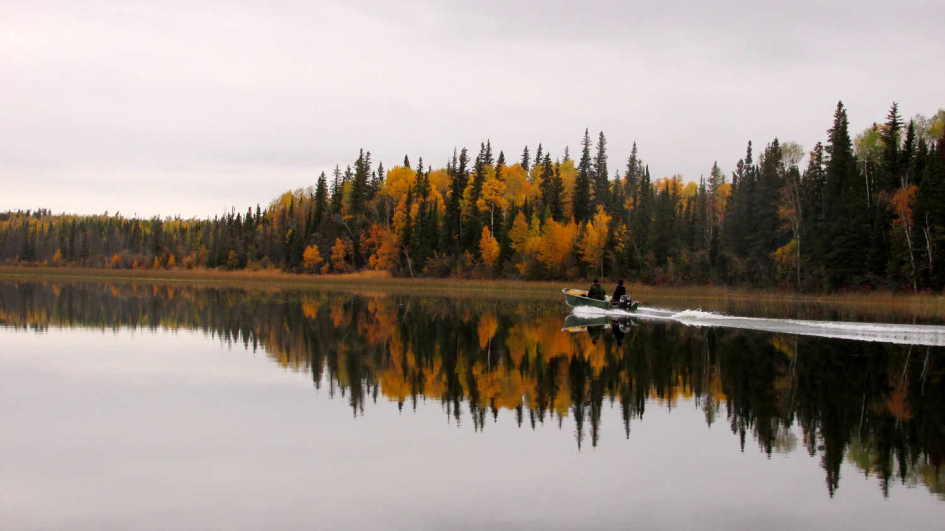 Two people travel by motorboat across a glassy lake that is connected to the Hayes River in the fall.