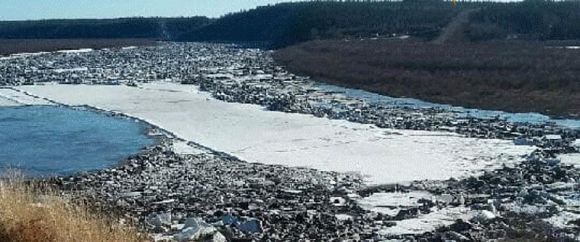 A high bank of the Arctic Red River taken from the town site across the river, with an arrow pointing to a trail head on the far hill. The river is in spring ice breakup mode. 