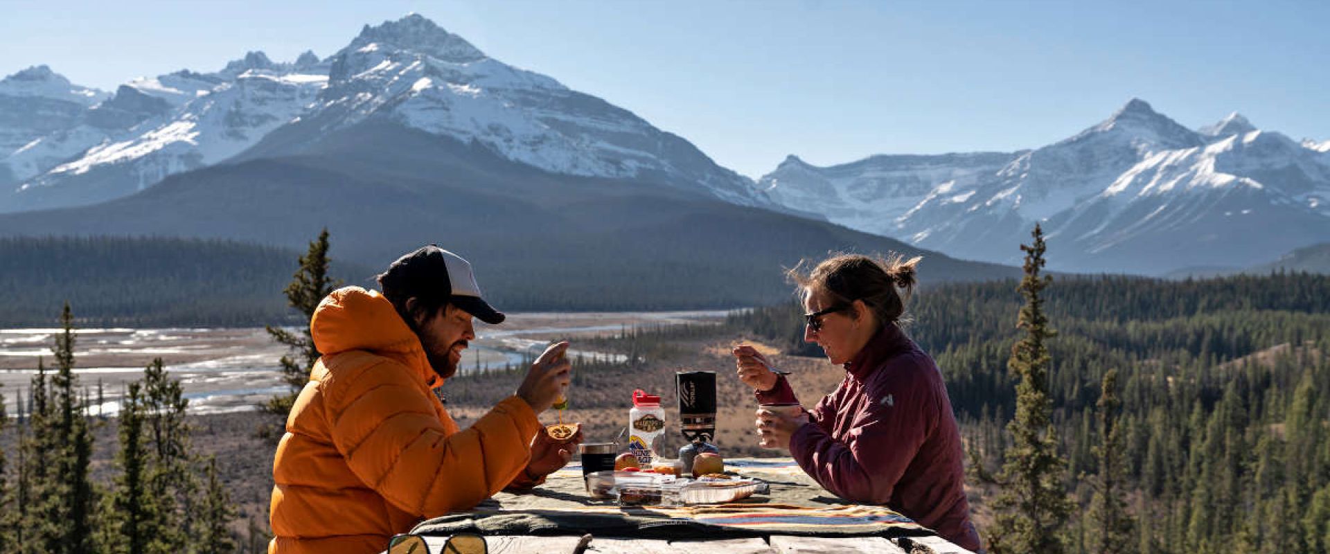 A couple sits at a picnic table at Howse Pass Viewpoint with backdrop of mountain peaks and the North Saskatchewan River.