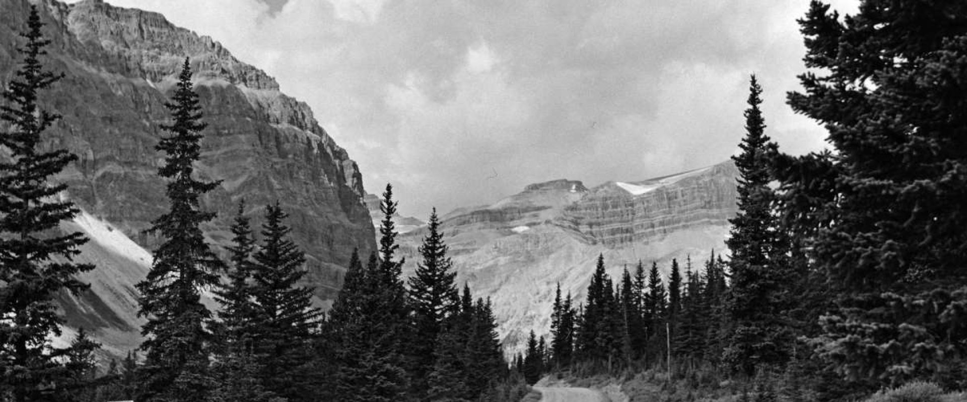 A black and white image of a car parked on the shoulder of the Icefields Parkway (a single lane gravel road) in 1949 with Crowfoot Glacier in the background.