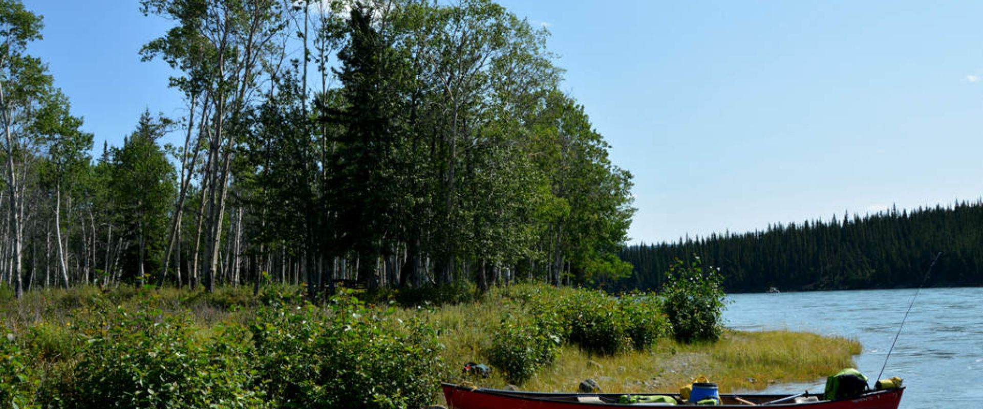 A beached canoe sits packed and ready on the grassy bank of the Thirty Mile River on a sunny day. 