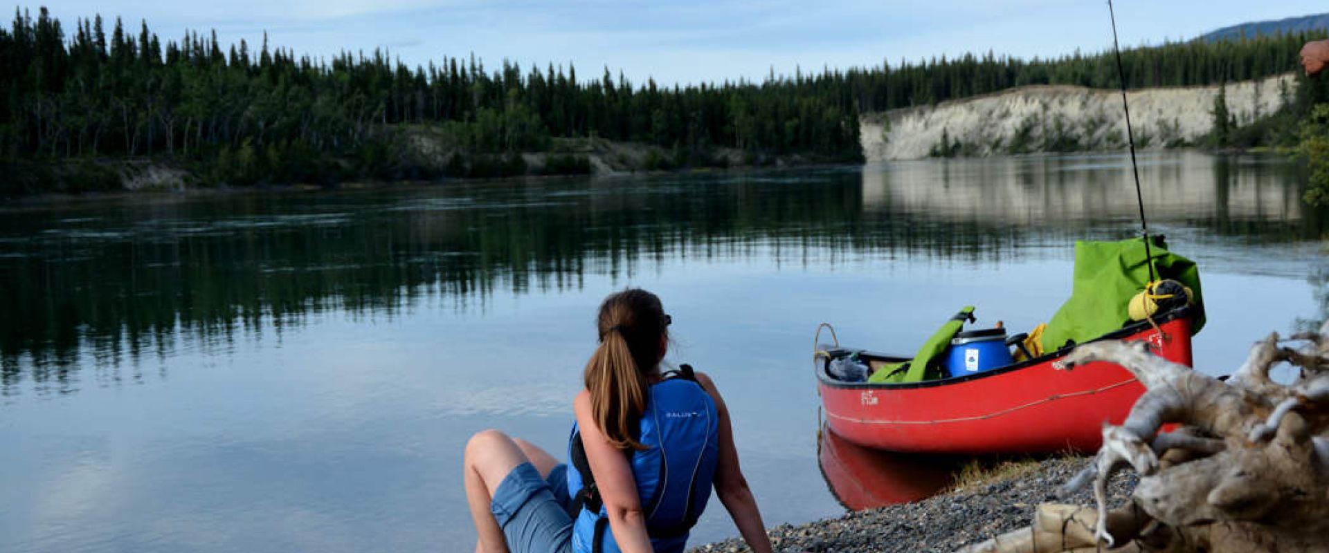 A woman sits next to her grounded canoe on the Thirty Mile River. The trees in the background are reflected onto the river.